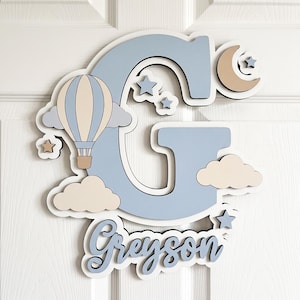 May include: A large, light blue wooden letter G featuring a hot air balloon, clouds, stars, and a crescent moon. Below the letter, the name Greyson is written in a blue cursive script. The design uses a layered, multi-dimensional aesthetic.