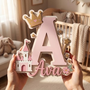 May include: A decorative wooden sign featuring a large light pink letter A with the name Ava written in cursive below. The sign is adorned with a gold crown, a pink and white castle, small gold stars, and a pink flower with green leaves.