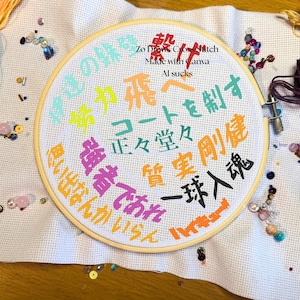 May include: A circular wooden embroidery hoop containing white Aida fabric with multi-colored Japanese kanji characters stitched in various fonts and colors. The text includes "Zo Draws Cross Stitch", "Made with Canva", and "AI sucks" at the top.