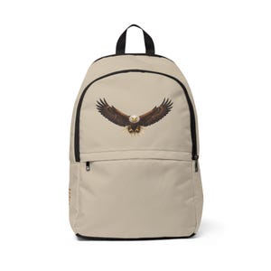 May include: A beige backpack featuring a graphic of a bald eagle with wings spread wide in flight. The backpack has a large main compartment, a smaller front zippered pocket, black trim, and a black top handle for carrying or hanging the bag.