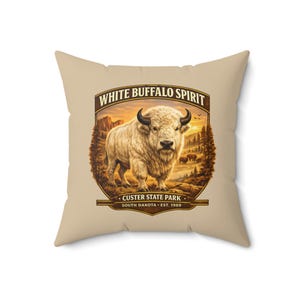 May include: A square beige throw pillow featuring a central graphic of a white buffalo standing in a landscape with trees and mountains. The text reads WHITE BUFFALO SPIRIT, CUSTER STATE PARK, SOUTH DAKOTA, EST. 1989 in a brown decorative frame.