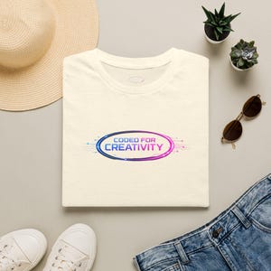 May include: A cream-coloured t-shirt featuring a central graphic of a blue and purple oval with circuit board lines and the text "CODED FOR CREATIVITY" printed in the centre. The shirt has a round neckline and a speckled fabric texture.