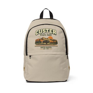 May include: A beige backpack with black trim and a top handle. The front features a graphic of Custer State Park, South Dakota, established 1889, depicting a bison, pine trees, mountains, and an orange sun against a light background.