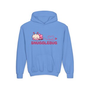 May include: A light blue hooded sweatshirt featuring a graphic of a pink cartoon bug with heart-shaped antennae. The bug is followed by a dotted heart trail and the red text SNUGGLEBUG is printed underneath the illustration on the chest.