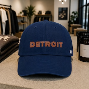 Puede incluir: Gorra de béisbol azul marino con la palabra "DETROIT" en letras naranjas en la parte delantera. La gorra tiene una visera curva y el número "47" bordado en el lateral. Hecha de tela suave.