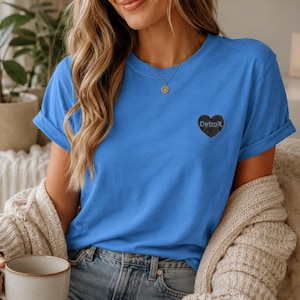 May include: A bright blue short-sleeved t-shirt featuring a small black heart graphic on the left chest. The heart contains the word Detroit written in a white, stylised font. The shirt has a crew neckline and rolled sleeves for a casual look.
