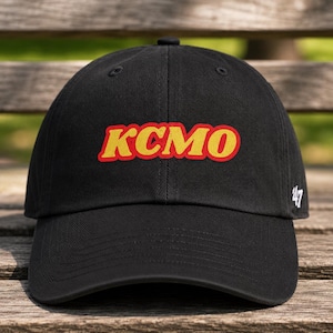 May include: Black baseball cap featuring the word "KCMO" in yellow with a red outline. The number "47" is embroidered on the side. The hat is displayed on a wooden surface.