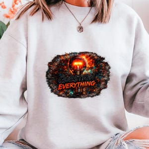 May include: A pale grey sweatshirt with a graphic of a lit lightbulb and the words "QUESTION EVERYTHING" in orange and white. The design has a dark, textured background with red, orange, and blue accents.