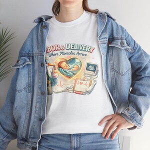 Op de afbeelding: Wit T-shirt met een kleurrijk grafisch ontwerp met de tekst "Labor & Delivery Where Miracles Arrive". Het ontwerp bevat een baby in een hart, een ooievaar en medische apparatuur.