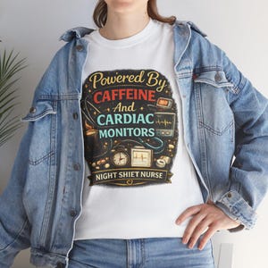Puede incluir: Camiseta blanca con un gráfico colorido que dice "Powered By Caffeine And Cardiac Monitors" con ilustraciones médicas y de café. El texto "Night Shift Nurse" está debajo del gráfico. La persona lleva una chaqueta vaquera azul.