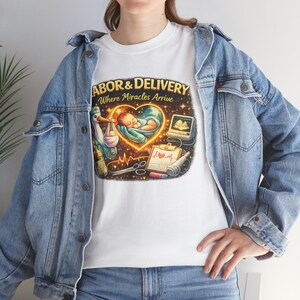Peut inclure: T-shirt blanc avec un motif graphique coloré. Le motif présente le texte "LABOR & DELIVERY Where Miracles Arrive" avec des illustrations d'un bébé, d'une cigogne et d'équipement médical. Le t-shirt est porté avec une veste en jean bleue.