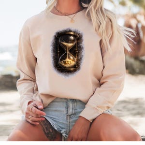 May include: A beige sweatshirt featuring a detailed hourglass design with the word "NEXT" at the bottom. The hourglass is set against a dark, textured background. The sweatshirt has long sleeves and a crew neck.