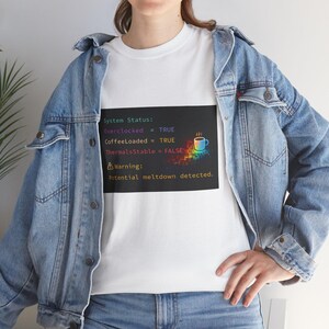 Puede incluir: Camiseta blanca con un gráfico que muestra un informe de estado del sistema. El informe incluye "Overclocked = TRUE", "CoffeeLoaded = TRUE" y "ThermalsStable = FALSE". Un mensaje de advertencia dice "Potential meltdown detected". También hay un gráfico de taza de café colorido.
