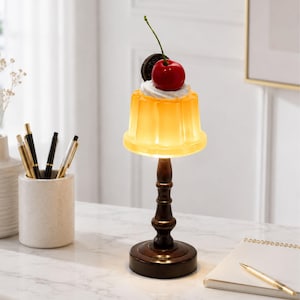 May include: A table lamp featuring a dark brown pedestal base and a translucent yellow jelly-shaped shade topped with a white cream swirl, a dark chocolate sandwich cookie, and a bright red cherry with a green stem, emitting a warm glow.
