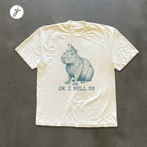 May include: White t-shirt featuring a teal illustration of a capybara with a bird on its head. The text "OK I PULL UP" is printed below the illustration. The shirt has short sleeves and a crew neck.