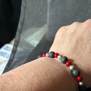 May include: A beaded bracelet featuring black lava rock beads, interspersed with red and silver-colored beads. The bracelet is worn on a wrist, showcasing the contrast of colors and textures.