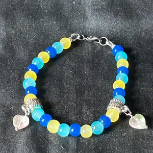 May include: A bracelet made of blue, yellow, and turquoise beads. The bracelet has silver-coloured accents and two heart-shaped charms. The beads are strung together with a silver-coloured clasp.