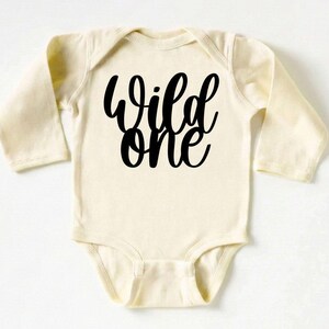 May include: Cream-colored baby bodysuit with long sleeves. The words "Wild one" are printed in bold, black script across the front. The bodysuit has a simple, classic design, suitable for a baby.