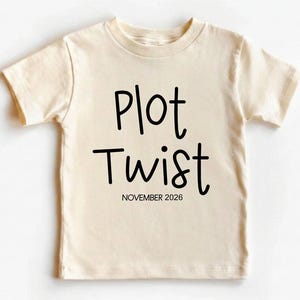 May include: A cream-colored toddler t-shirt with the words "Plot Twist" in black script. Below the text is the date "NOVEMBER 2026". The shirt is made of soft material and has short sleeves.