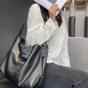 May include: Black shoulder bag with a silver emblem. The bag has a long strap and a buckle closure. The bag is made of a textured material. The bag is a tote style.