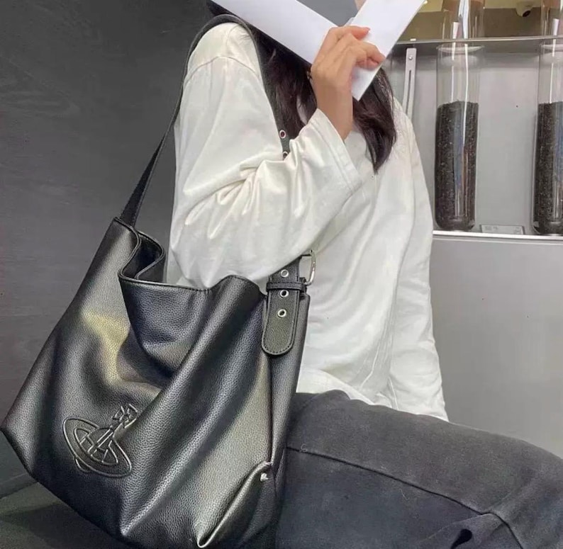 May include: Black shoulder bag with a silver emblem. The bag has a long strap and a buckle closure. The bag is made of a textured material. The bag is a tote style.