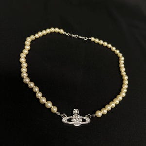 May include: Cream pearl necklace with a silver pendant. The pendant has a detailed design with clear crystals. The necklace is shown against a black backdrop.