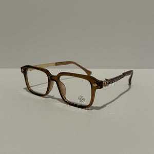 May include: Brown rectangular eyeglasses with gold-toned arms and decorative detailing on the temples. The lenses are clear, and a small emblem is visible on the lower right lens. The glasses are set against a neutral background.