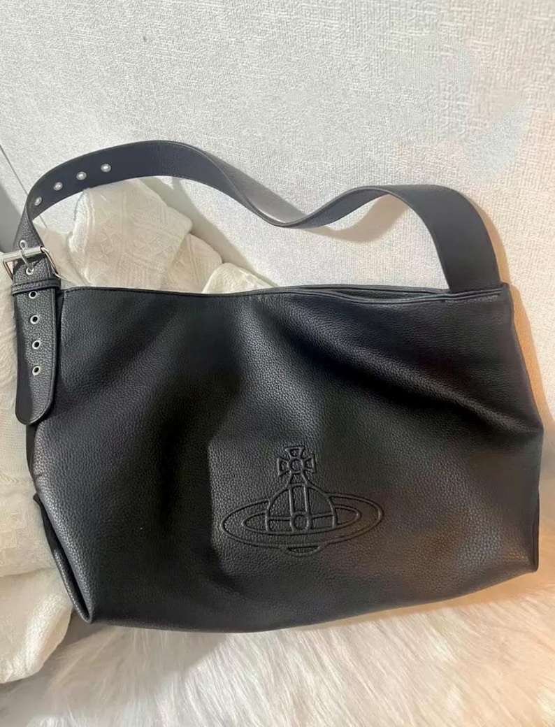 May include: Black leather shoulder bag with a large strap and a silver buckle. The bag features a debossed emblem on the front. The bag has a soft, textured appearance and a long, adjustable strap.