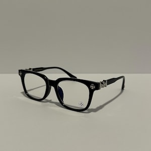 May include: Black eyeglasses with a rectangular frame. The temples and corners feature silver-colored decorative accents. The lenses are clear, and the frame has a small, decorative emblem on the lower corner.