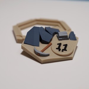Op de afbeelding: Een beige zeshoekig spelstuk met een pikhouweel, blauwe rotsformaties en het nummer 11. Op de achtergrond een groter, beige zeshoekig frame. Waarschijnlijk een onderdeel van een bordspel.