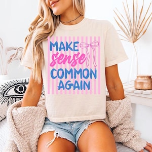 Może przedstawiać: Kremowy t-shirt z napisem "MAKE sense COMMON AGAIN" w kolorze niebieskim i różowym, na różowym, pasiastym tle. Koszulka ma krótkie rękawy i luźny krój.