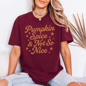 Könnte beinhalten: Ein kastanienbraunes Kurzarm-T-Shirt mit dem goldenen Schriftzug Pumpkin Spice & Not So Nice auf der Vorderseite. Das Design enthält mehrere kleine goldene Sterne, die um den in Schreib- und Serifenschrift gehaltenen Text verteilt sind.