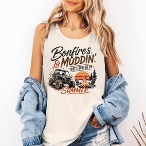 May include: A cream-coloured sleeveless tank top featuring a graphic print with the text Bonfires & MUDDIN' THAT'S HOW WE DO SUMMER. The design includes an off-road vehicle, a campfire, two chairs, and a sunset set against a backdrop of pine trees.