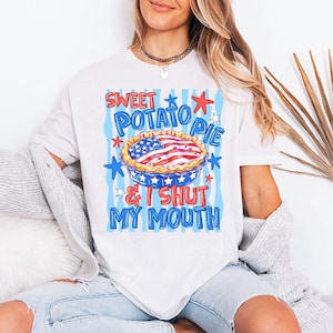 Op de afbeelding: Een wit t-shirt met korte mouwen en een grafische print van een taart met een Amerikaans vlagpatroon. De tekst SWEET POTATO PIE & I SHUT MY MOUTH staat in blauwe en rode letters, omringd door sterren op een lichtblauwe achtergrond met strepen.