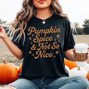 Könnte beinhalten: Ein schwarzes Kurzarm-T-Shirt mit dem orangefarbenen Text Pumpkin Spice & Not So Nice, verziert mit kleinen orangefarbenen Sternen. Das T-Shirt hat einen Rundhalsausschnitt und wird zu einer blauen Jeans und einem weißen Kaffeebecher getragen.