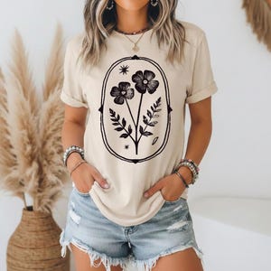 May include: A beige short-sleeved t-shirt featuring a black graphic print of two flowers and fern-like leaves enclosed within an oval frame. The design includes small star accents above and below the floral arrangement, centered on the chest.