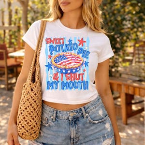 Op de afbeelding: Een wit T-shirt met korte mouwen en een afbeelding van een taart met een Amerikaans vlagpatroon op de korst. De tekst "SWEET POTATO PIE & I SHUT MY MOUTH" staat in blauwe letters, omringd door rode en blauwe sterren en blauwe strepen.