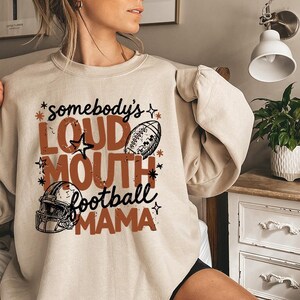 Könnte beinhalten: Ein beigefarbenes Sweatshirt mit Rundhalsausschnitt und einem Grafikdruck mit dem Text "somebody's LOUD MOUTH football MAMA" in brauner und schwarzer Schrift. Das Design enthält Illustrationen eines Footballs und eines Helms, akzentuiert durch kleine Sterne.
