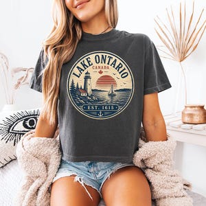 May include: A charcoal gray short-sleeve t-shirt featuring a circular graphic print. The design depicts a lighthouse, a sailboat, and a sunset over Lake Ontario, Canada, with the text "LAKE ONTARIO CANADA EST. 1615" and an anchor icon.