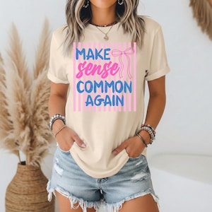 Może przedstawiać: Kremowy t-shirt z napisem "MAKE sense COMMON AGAIN" w kolorze niebieskim i różowym. Tekst umieszczono na różowym, pasiastym tle z różową kokardą. Koszulka ma krótkie rękawy i luźny krój.