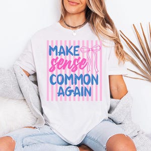 Op de afbeelding: Wit T-shirt met de tekst "MAKE sense COMMON AGAIN" in blauw en roze, met een roze strik. De tekst staat tegen een achtergrond van roze en witte verticale strepen. Het shirt is een casual stijl.