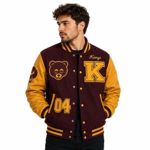 Può includere: Una giacca varsity marrone con maniche in pelle gialla, caratterizzata da una grafica a testa d'orso gialla, la lettera K, il nome Kanye e il numero 04 ricamati in giallo. La giacca presenta una chiusura frontale con bottoni e bordi a righe.
