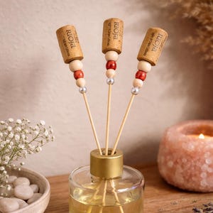 Könnte beinhalten: Ein klarer Glas-Diffusor für ätherische Öle mit goldfarbenem Deckel. Der Diffusor enthält Holzstäbchen und ist mit Holzperlen, roten Perlen und silbernen Perlen verziert. Die Stäbchen sind mit Korken mit der Aufschrift "Rioja" versehen.