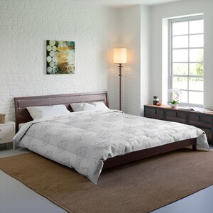 May include: A white comforter featuring a repeating black geometric diamond pattern, draped over a dark brown wooden bed frame with a horizontal slat headboard, accompanied by two white pillows in a bedroom setting with a floor lamp.