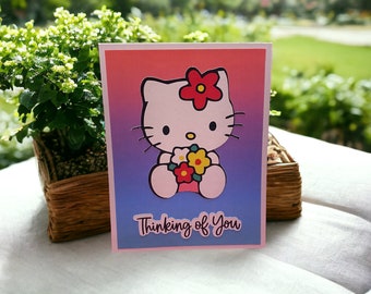 以下が含まれることがあります： 赤い花をつけた漫画の猫のキャラクターが、花のブーケを持っているグリーティングカード。カードは、ピンク、青、紫のグラデーションの背景に「Thinking of You」の文字が入っています。