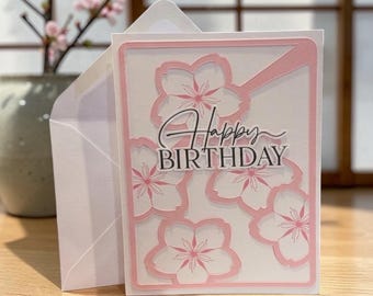 May include: A white rectangular greeting card featuring a layered pink die-cut frame and several pink cherry blossom silhouettes. The center of the card displays the black text Happy BIRTHDAY in a combination of script and serif fonts.