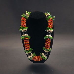 May include: A vibrant lei necklace featuring pineapple-shaped elements in shades of orange and red, accented with green leaves and white flowers. Black beads are interspersed throughout the design. The necklace is displayed on a black mannequin.