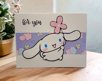 以下が含まれることがあります： 白いカードに黒い筆記体で「for you」と書かれています。ピンクの花を頭に乗せた漫画の犬が花を持っています。犬はピンクと白の花が咲いた紫色の背景の上にいます。