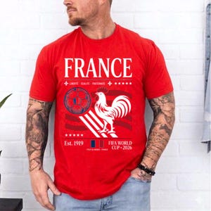 May include: A red short-sleeved t-shirt featuring the word FRANCE in white text, a graphic of a soccer ball, a white rooster, and the text LIBERTE EGALITE FRATERNITE, Est. 1919, and FIFA WORLD CUP 2026 printed in white on the front chest area.