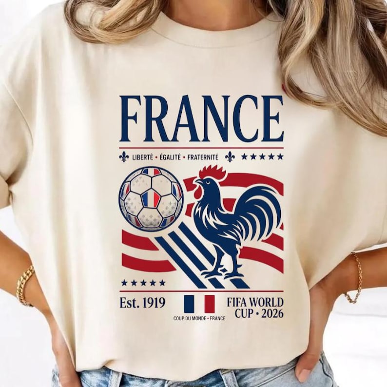 May include: A cream-colored t-shirt featuring a graphic with the word FRANCE in large blue letters. The design includes a soccer ball, a blue rooster, red and blue stripes, the text LIBERT&Eacute; &Eacute;GALIT&Eacute; FRATERNIT&Eacute;, Est. 1919, and FIFA WORLD CUP 2026.